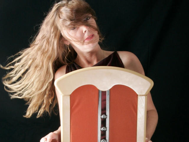 Simone-69 with long, messy blonde hair holding a wooden chair with red fabric and metal buttons, black sleeveless top, black background, looking up with slightly parted lips.