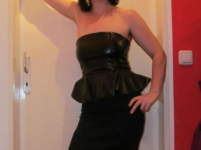 Wetgerman-hot in a black strapless leather top with a ruffle and a black skirt, standing with one hand on her hip, showing her tits and ass.