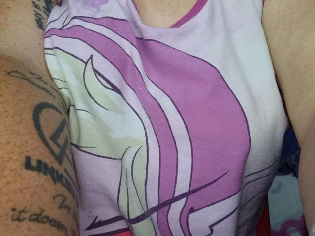 Wetgerman-hot's pink and purple tank top with a stylized design, showing their tits, with a tattooed arm on the left. Skin is light, tattoos are black.