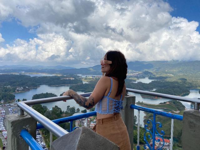 Sara Duncan, con tatuaggi, indossa un reggiseno di pizzo blu e una gonna color carne, mentre guarda indietro da un balcone, con il sedere leggermente visibile, sotto un cielo nuvoloso..