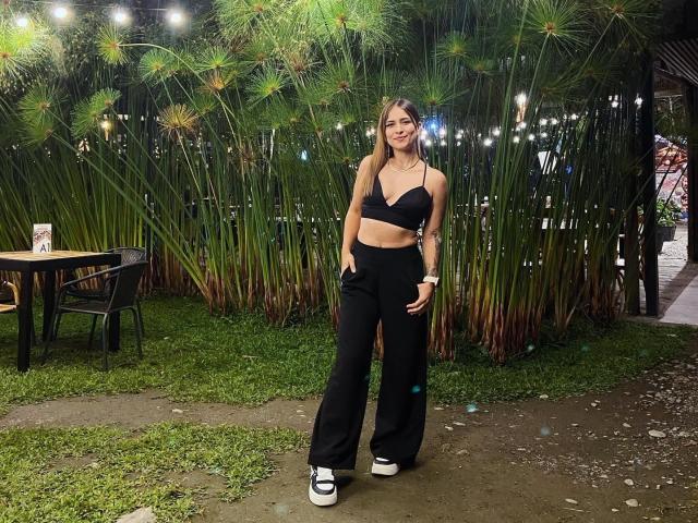 SaraDuncan with long blonde hair, black crop top, black high-waisted pants, and white sneakers, standing in a grassy outdoor area, hands in pockets, smiling.