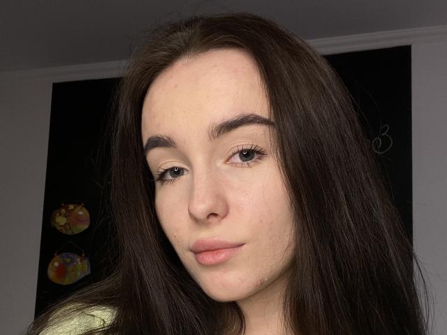 Milana Furst tiene el pelo largo, castaño oscuro, piel clara y un maquillaje discreto con labios rosados. Sus cejas son oscuras y tiene pechos pequeños y lindos. Mira directamente a la cámara..