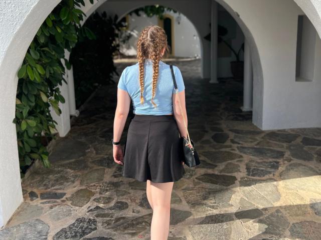 EllyNora-hot with long braided hair, light blue top, black skirt, holding black bag, standing on stone path under white archway, wearing black wristband.