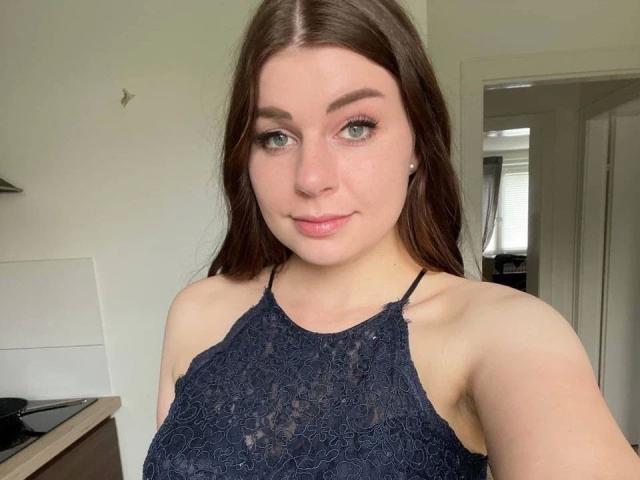 EllyNora-hot with long brown hair, green eyes, and a small nose, wearing a dark lace halter top, showing her bare shoulders and small tits. She's smiling slightly.
