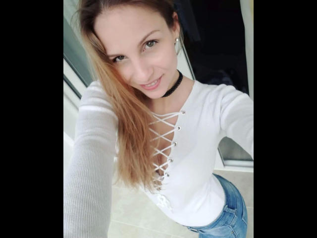 MaryWet-hot with long blonde hair, white lace-up top, choker, and blue jeans, smiling at the camera, showing her tits and cute face, standing in a bright room.