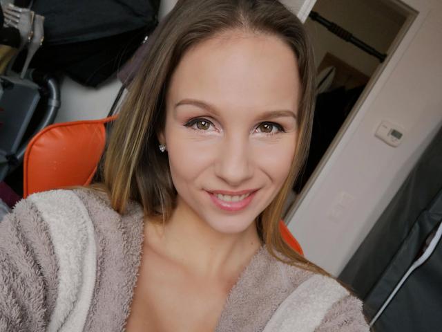 MaryWet-hot, smiling with green eyes, wearing a fluffy beige and white robe, has straight brown hair, small earrings, and a cute face, sitting on an orange chair.