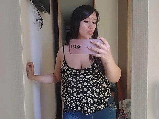 LizBellaa with long black hair, wearing a black floral top, showing small tits, and blue jeans, taking a mirror selfie with a pink phone, pouting her lips.