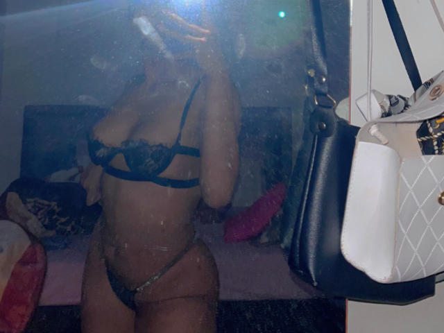 Stephanie Davies trägt einen schwarzen Spitzen-BH und String, zeigt ihre Brüste und ihren Hintern und macht ein Selfie vor einem Spiegel mit einem Taschenlampe. Im Hintergrund sind eine schwarze Handtasche und eine weiße Tasche zu sehen..