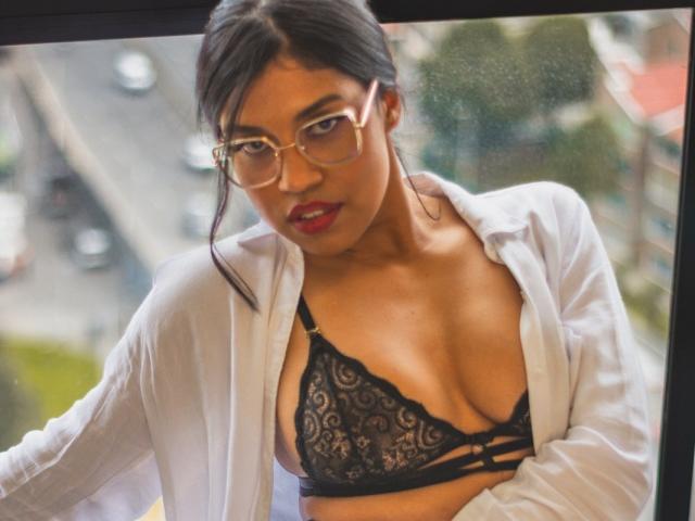 Stephanie Davies mit Brille, rotem Lippenstift und einem schwarzen Spitzen-BH, trägt ein offenes weißes Hemd und blickt durch ein Fenster. Dabei sind ihre Brüste zu sehen, und sie hat einen sexy und selbstbewussten Ausdruck..