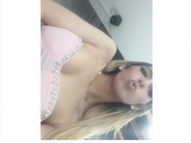 Putinna with blonde hair, pouty lips, wearing a pink strapless top with white pearl trim, showing her small tits, wearing pearl earrings, and a gold necklace.