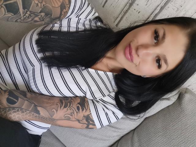 KiraKane, attraente, con capelli neri, una camicia a righe, tatuaggi sulle braccia e labbra rosa, seduta su un divano grigio, che guarda la telecamera con un leggero sorriso..