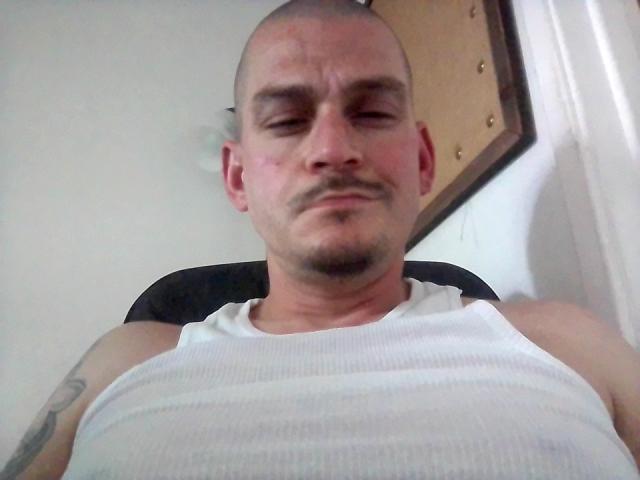 MrAprils with a bald head, slight mustache, and white tank top, showing a pensive expression, visible tattoo on left arm, sitting in a black chair.