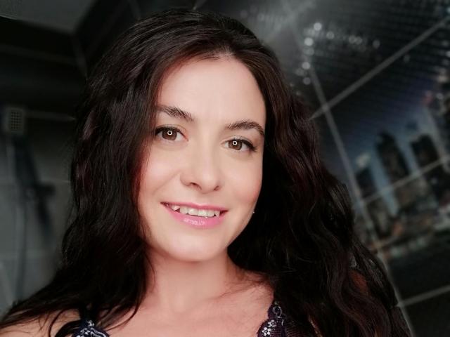 CindyStrawberry-hot has dark brown wavy hair, brown eyes, pink lipstick, and a cute smile, wearing a sparkly purple top. She looks attractive and is smiling directly at the camera.