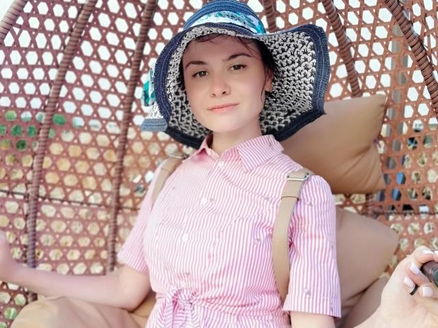CindyStrawberry-hot with pale skin, small nose, and brown eyes, wearing a pink striped shirt and blue straw hat, sitting in a wicker chair with beige cushions.