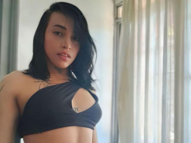 YeiSela, avec ses cheveux noirs, portant un haut noir découpé, laissant entrevoir de petites seins, avec des tatouages sur la poitrine et le haut du bras, se tient près d'une fenêtre avec des rideaux transparents..