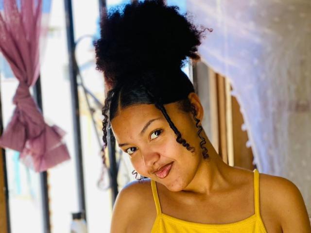 Sirina with dark, curly hair in a high bun, wearing a yellow tank top, smiling slightly at the camera, showing a cute, confident, and attractive face.