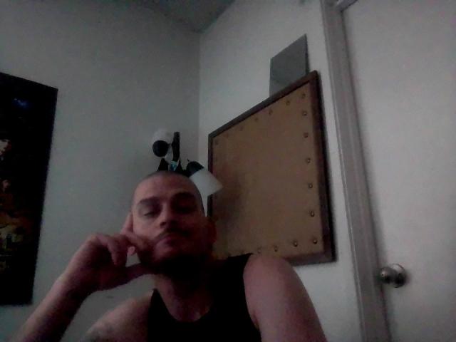 O Sr. Aprils tem a cabeça careca com uma câmera montada em cima, veste uma regata preta e está falando ao telefone com uma expressão séria. Ele está em um quarto com pouca luz..