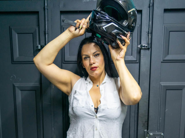 LizaRussell with medium tits, black hair, and red lipstick, wearing a white sleeveless shirt, holding a black motorcycle helmet above her head, arms raised.