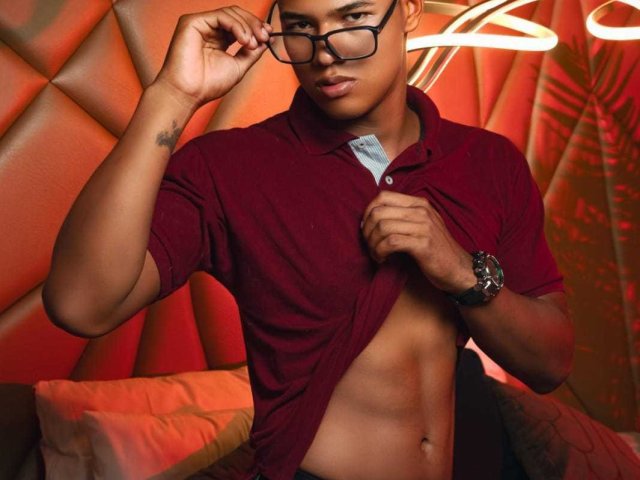 DarreIHot, a sexy brown dude with glasses, lifting a red polo to show his toned abs, wearing a black watch, tattoo on left arm, in a red-lit room.