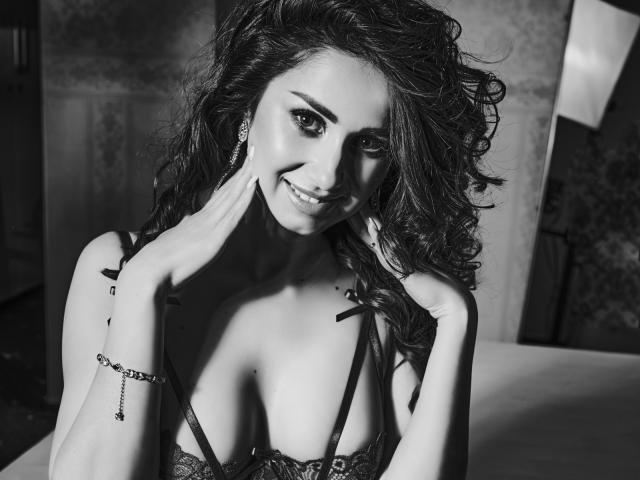 Black-and-white photo of SelenaWest with curly hair, smiling, hands near face, wearing lace bra, showing small tits, bracelet on left wrist, earrings, and slight cleavage.
