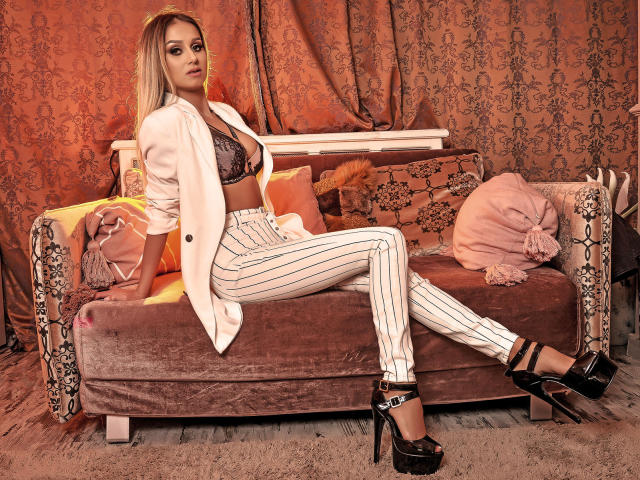 SelenaWest with blonde hair, wearing a pink blazer, striped white pants, black high heels, and a lace bra, sitting on a brown velvet sofa, showing her ass.