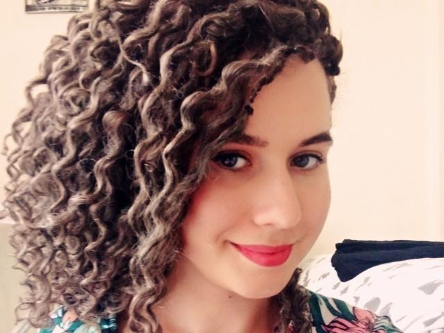 SelenaXCute, con i capelli castani ricci, il rossetto rosso e un leggero sorriso, indossa una maglietta colorata, guarda la fotocamera con gli occhi azzurri, mostrando un viso carino e un bel sedere..