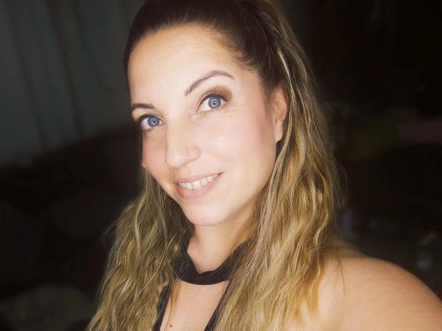 SexyStine-hot has blue eyes, wavy blonde hair, and a smile, wearing a black halter top, with fair skin and a cute nose, in a dimly lit setting.