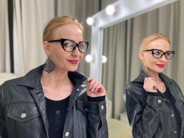 SquirtyAlisa-hot with blonde hair, black glasses, red lipstick, black leather jacket, black top, and large ornate earrings, applying makeup in front of a mirror. Reflection visible.