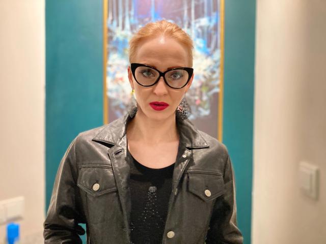 SquirtyAlisa-hot with red lipstick, black glasses, and black leather jacket, wearing a black top, standing in a room, blue-green wall in background, hands by sides, looking forward.