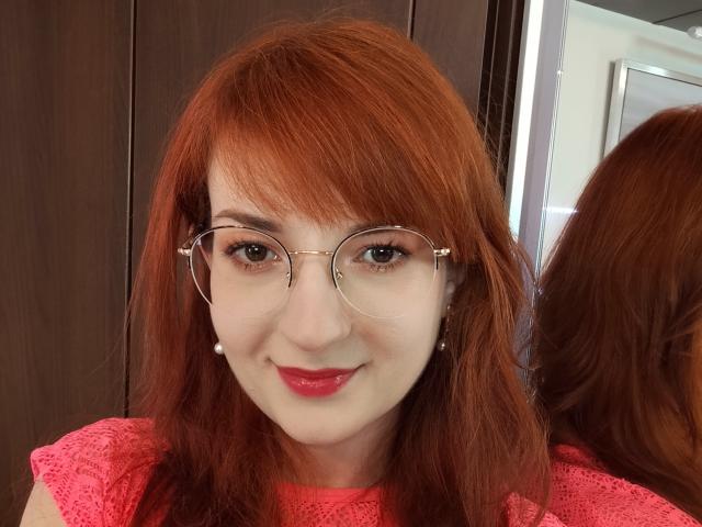 MoonBlossom with red hair, glasses, and pink lace top, showing off her cute face and red lipstick, smiling at the camera, with her reflection in the mirror.