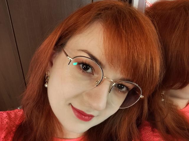 MoonBlossom has red hair, glasses with thin metal frames, and red lipstick. She's smiling with fair skin, small nose, and pearl earrings. Her ass is not visible.