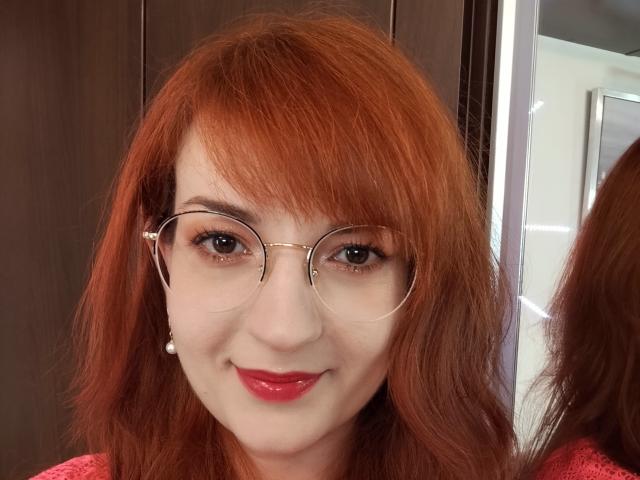 MoonBlossom has red hair, glasses, and is smiling with red lipstick. She's wearing a pearl earring and a pink top. Her tits are not visible.