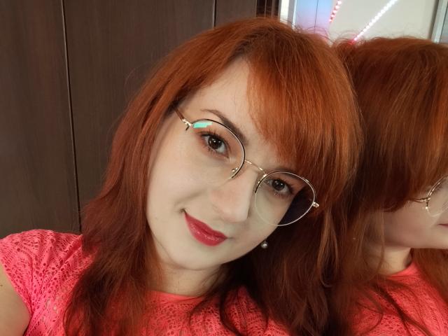 MoonBlossom with red hair, glasses, and pink lace shirt, smiling at the camera, reflecting in the mirror. Her red lips and fair skin are visible.