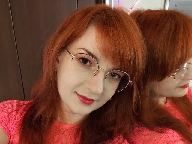 MoonBlossom with red hair, glasses, and pink lace shirt, smiling at her reflection. She's got cute tits and a soft, fair complexion. Her red lipstick matches her hair.