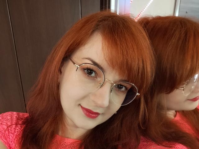 MoonBlossom has red hair, glasses, and a pink lace shirt, with a reflection in the mirror showing her smiling face and red lipstick. Her tits are not visible.