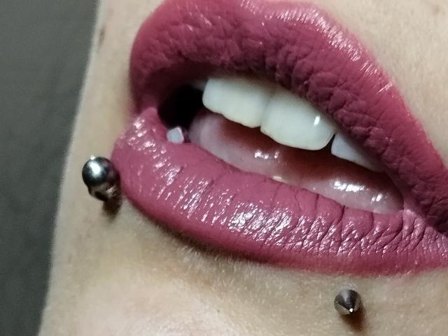 RivieraM's glossy pink lips with two silver lip piercings on the lower left and right side, showing white teeth and a slightly open mouth.