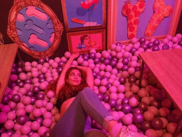 KendallPiierce69 with curly red hair, blue jeans, and white sneakers, lying in a purple and white ball pit, arms raised, looking up, in a brightly colored, playful room.
