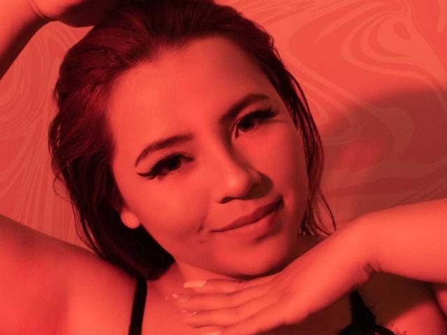 MelisaBrunette with red-tinted skin, dark hair, and eyeliner, smiling slightly, hands near her face, black strap visible, looking at camera. Red light casts warm glow.
