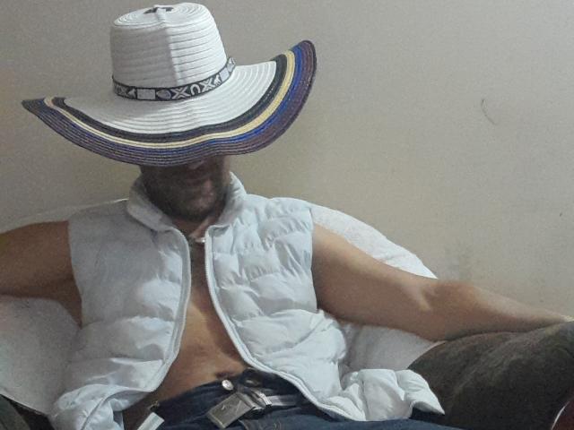 Arsoleros is wearing a white, striped, wide-brimmed hat with blue and yellow trim, a sleeveless puffy vest, and no shirt, showing a bare, hairy chest.