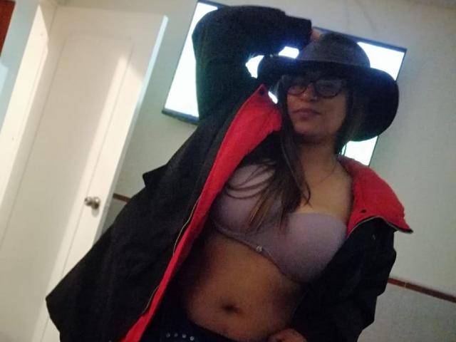 TompsonKarina in a black jacket with red lining, wearing glasses and a hat, showing her tits in a pink bra, with her hand on her head, and a slight smile.