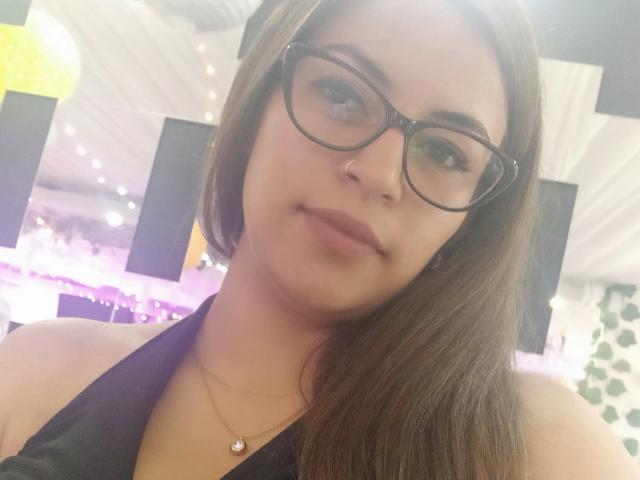 TompsonKarina has brown hair, glasses, a nose ring, and a subtle smile. She's wearing a black halter top and a small necklace with a heart pendant.
