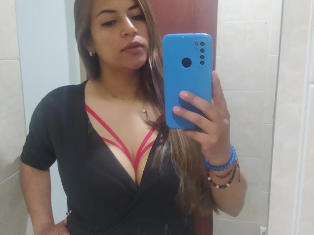 TompsonKarina with long brown hair, black top, and red bra straps, showing her tits in a mirror selfie with a blue phone, wearing bracelets and a necklace.