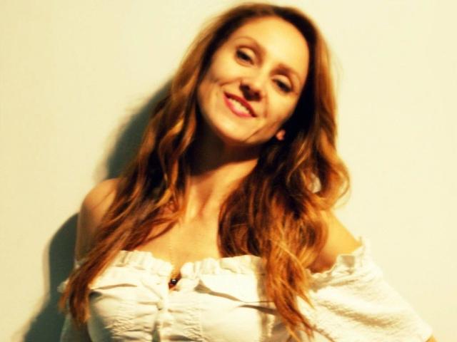 LarisaRais-hot with wavy brown hair, smiling, wearing an off-shoulder white top, exposing her tits, against a plain background, looking slightly blurred.