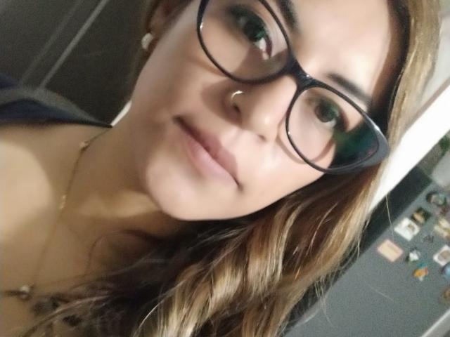 TompsonKarina with glasses, nose ring, and wavy brown hair, smiling softly. She's got cute tits, subtle makeup, and a gold necklace. Her green eyes are focused.