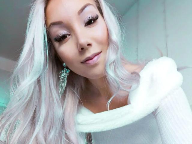 Lady-Khaleesi with long, wavy white hair, wearing a white fur-collared top, and dangling earrings, has a cute smile and long eyelashes, looking down at the camera.