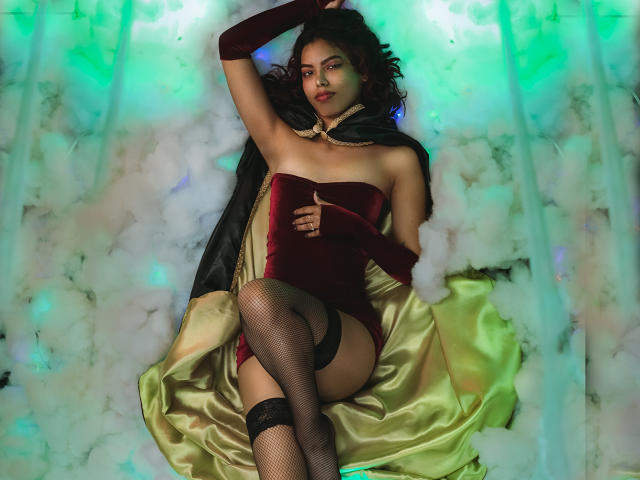 RouseGilmore with dark hair, red velvet strapless top, black fishnet stockings, and gold-green satin skirt, laying on fluffy white, green-blue lit background, wearing black gloves.