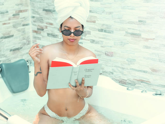 RouseGilmore with towel on head, sunglasses, and white lace panties, reading a book in a bathtub, water up to her thighs, holding the book with one hand.