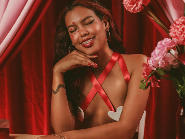 RouseGilmore, con i capelli rossi e ricci, indossa una maglietta con un fiocco rosso, ha dei piccoli copriceti a forma di cuore che coprono i seni, ha le unghie rosse, sta sorridendo, toccandosi il mento, ci sono fiori rosa, tende rosse e un tatuaggio di un serpente sul polso destro..