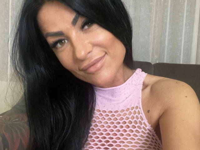 AndraD has long black hair, dark eyebrows, and is smiling slightly. She's wearing a pink mesh top that reveals her small tits. Her skin is tan.