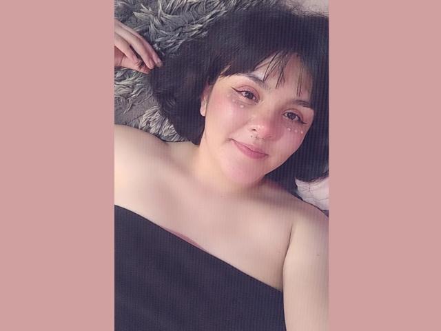 SofiaParker69 with short black hair, light skin, small nose ring, and eyeliner, lying on a fluffy grey surface, topless, black cloth covering her tits. Soft pink background.